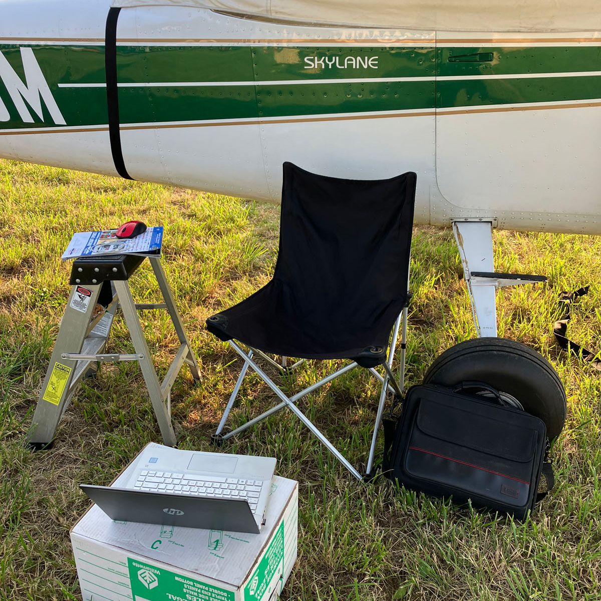 camp set up with laptop, laptop on box, folding chair, and step ladder under wing of plane